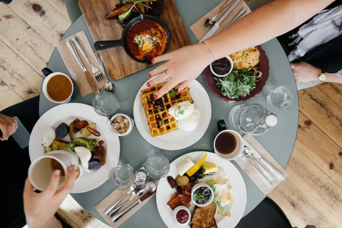 Overhead view of a shared brunch table — waffles, eggs, coffee, and a hand reaching in for food